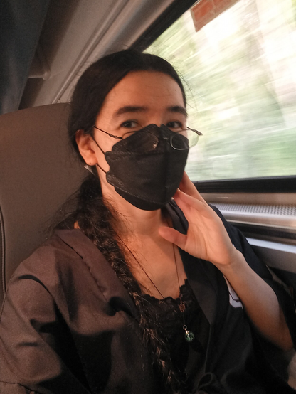 A woman in a black mask and glasses with long, braided black hair sitting in a train. she wears a black, lacy camisole under a black haori with a necklace with a green orb hanging down her chest. She has her hand raised to the cheek, looking over her glasses at the camera with her black eyes. The faintest sign of crow's feet can be seen at the corners of her eyes, hinting at a smile.