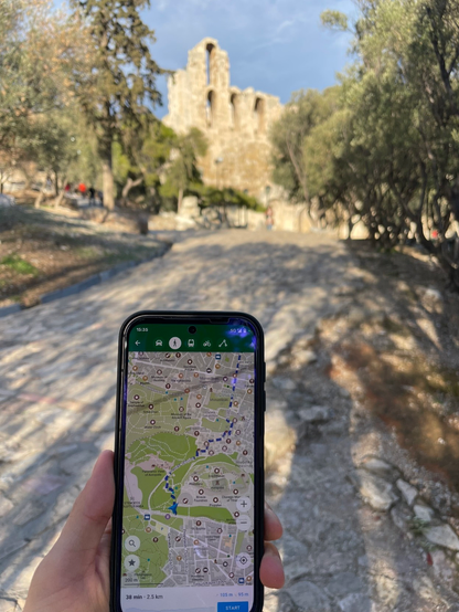 A picture of my hand holding my Fairpohone 5 with Organic Maps in navigation mode, and Ancient Greece ruins in the background.