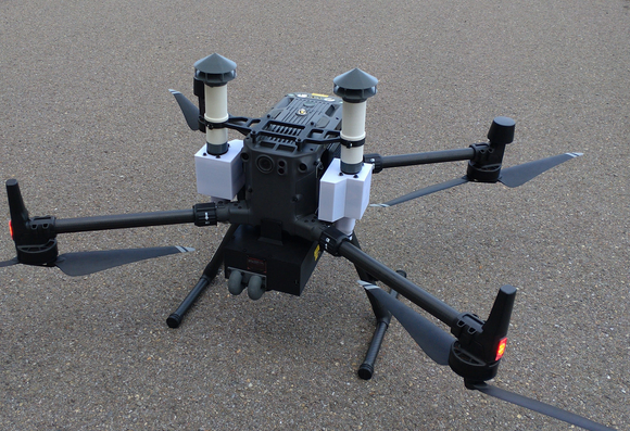 A dji Matrice M300 multicopter on asphalt with 3d printed parts attached all over, two "chimineys" at either side for particle measurements and a black "belly" underneath, with temperature, humidity and gas sensors.
