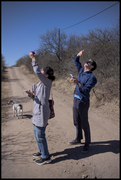 two people standing on a dirt road, looking up into the sky, shielding their eyes from the sun with a hand each. the left person carries a mate in the hand they use to shield their eyes. behind them is a dog looking around