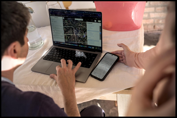 an over the shoulder view between two people who sitting/standing around a table and look at a laptop and phone that rest on the table. the laptop shows an openstreetmap editor open