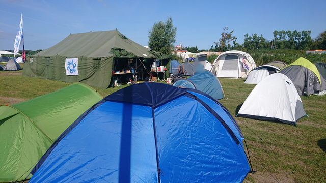 The sunny campsite, showing the NixOS village, including a large army tent flying a NixOS flag, smaller tents around it, and some greenery in the background.