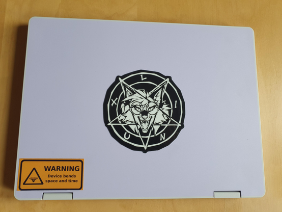 A Framework 12 laptop in lavender colour scheme, shown closed from the top. The middle of the laptop, where the Framework logo normally is, has been covered with Neotheta's black-and-white Xenia "pentacle" sticker. In the bottom left corner, a yellow sticker says "WARNING: Device bends space and time".